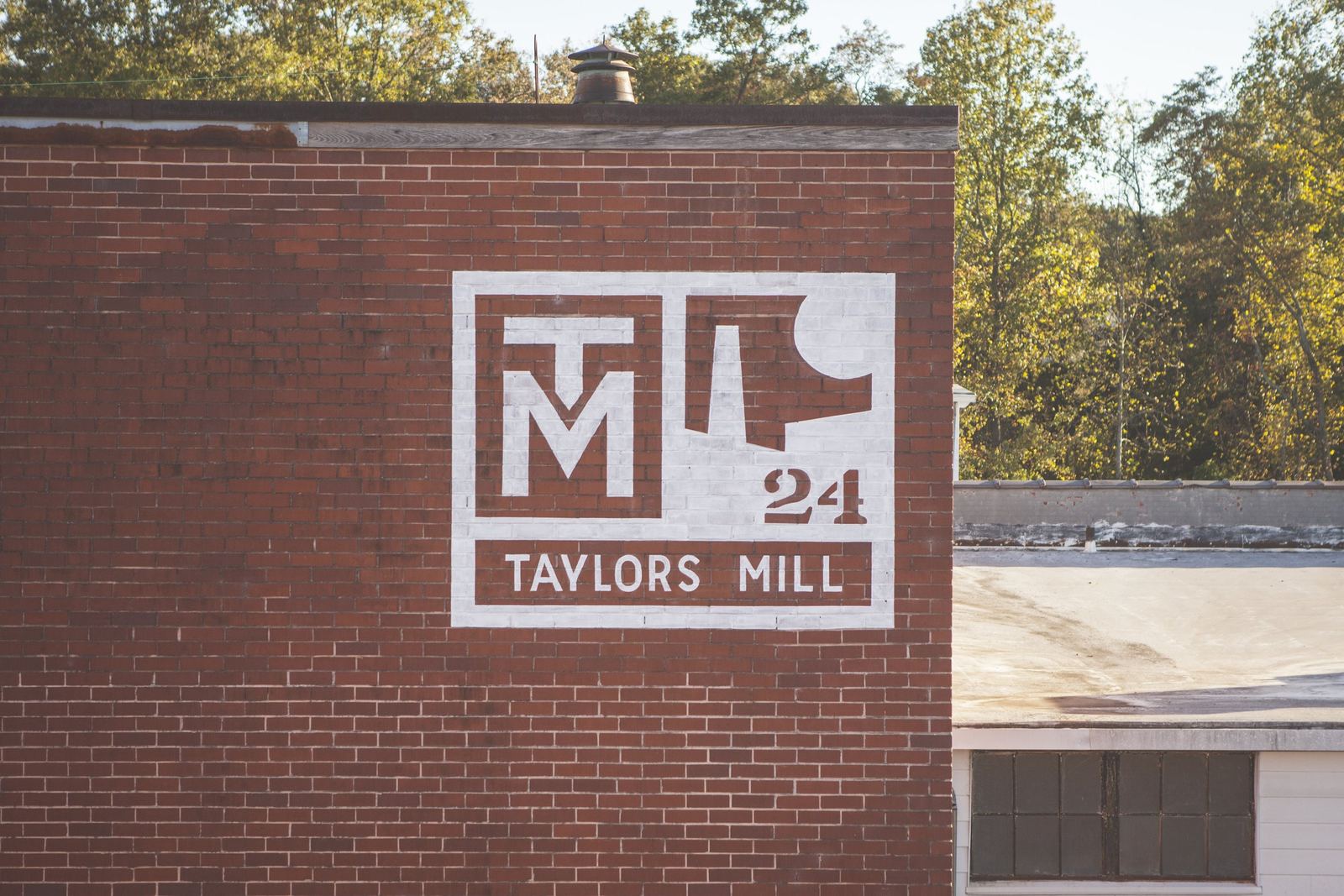 Brick wall featuring a painted sign that reads "Taylors Mill 24" with a logo and the letters M and T.