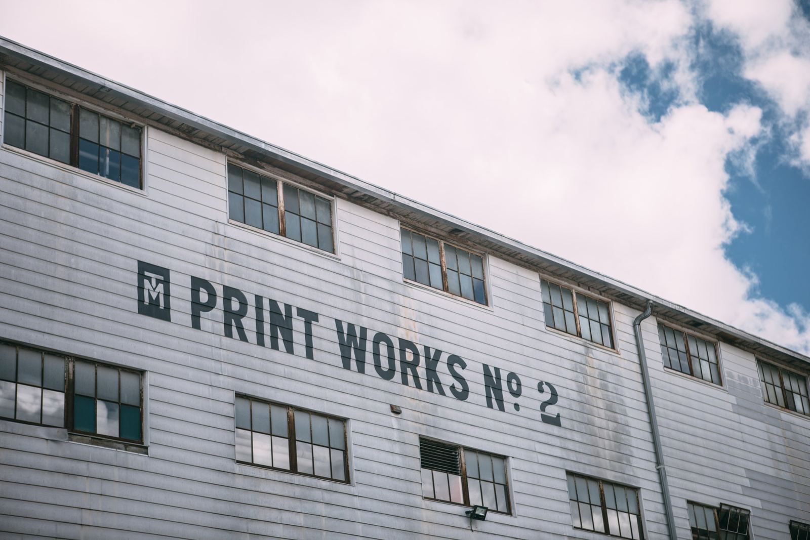 A white clapboard building with rows of windows and large black lettering that reads “Print Works No. 2” beside a boxed T M logo, under a partly cloudy sky.