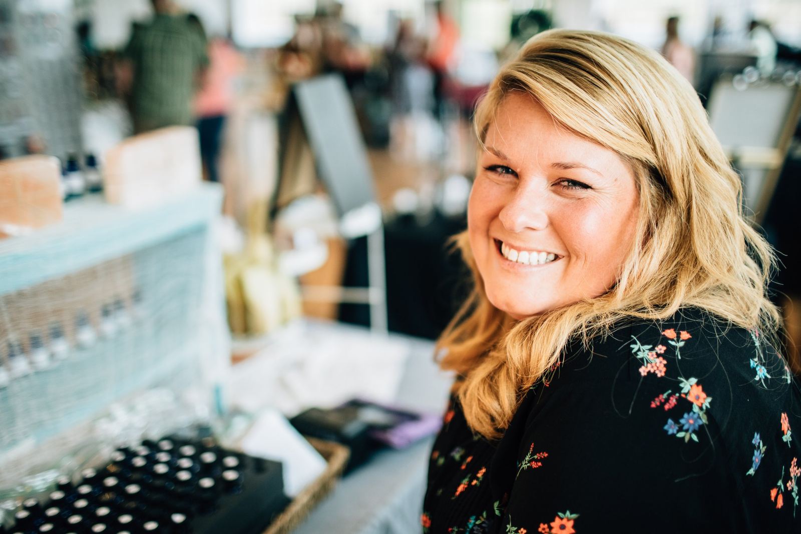 A smiling woman with blonde hair at an indoor market or craft fair, wearing a black top with floral patterns, with various items displayed on a table nearby.