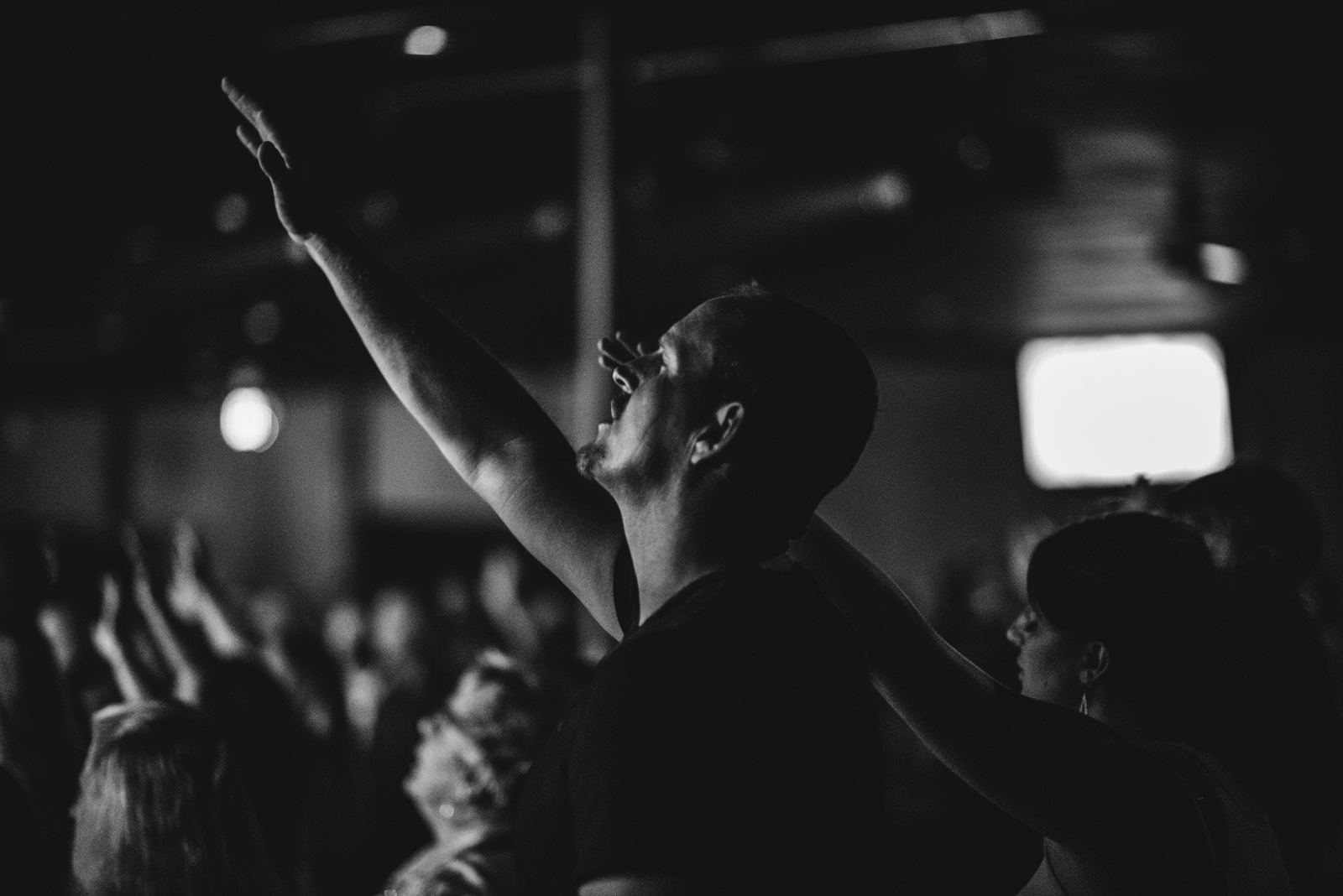 A black-and-white photo of a man in a dim room with one arm raised and his head tilted back, with other raised hands blurred behind him.